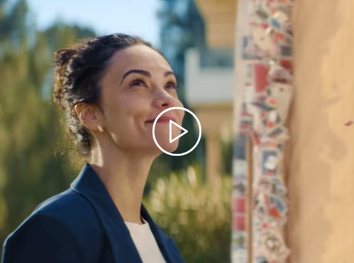 Cette image est une capture de la campagne LA BANQUE POSTALE dans laquelle nous voyons une femme ajouter la carte manquante au chateau de carte. Sur cette carte, on peut y voir le logo de la Banque Postale. Son geste représente son rôle de conseillère et plus largement le rôle de la banque postale dans la concrétisation et la consolidation des projets de chacun, afin qu'un château de carte, fragile, puisse devenir une maison, solide.